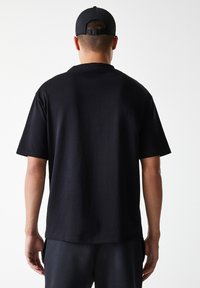 Black short-sleeve t-shirt made of cotton, featuring a round neckline and straight cut. Worn with a black cap, it has a smooth texture.