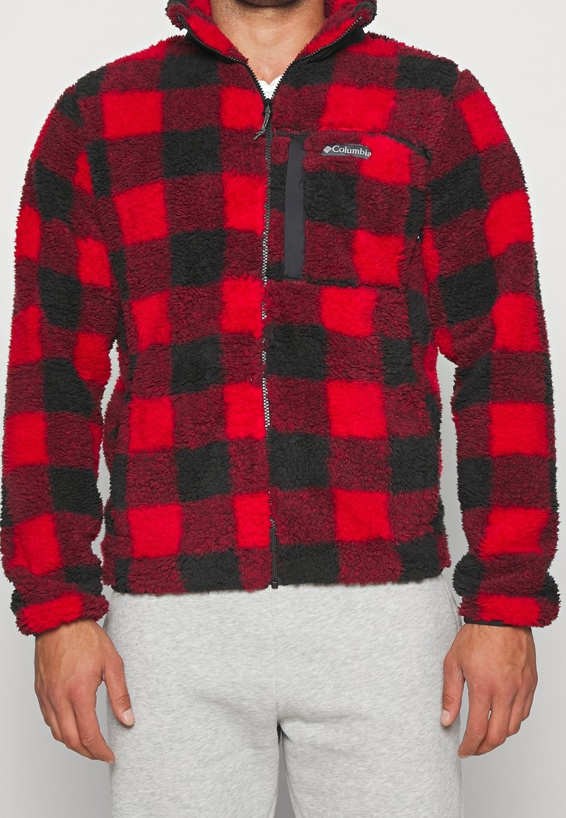 Homme portant une veste polaire à carreaux rouges et noirs avec fermeture éclair et poche poitrine, associée à un pantalon gris clair.