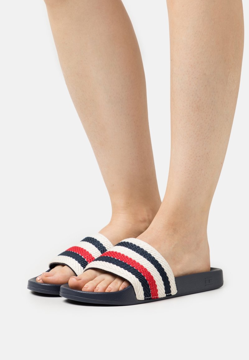 Pieds portant des sandales à enfiler bleu marine avec des brides blanches tissées comportant des rayures horizontales rouges et bleu marine, sur un fond uni clair.