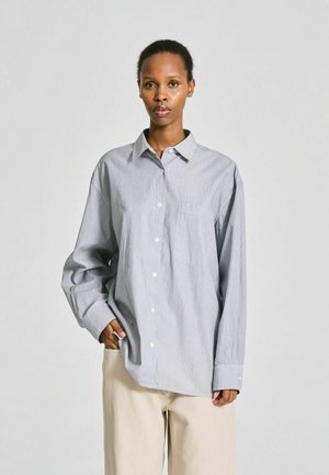 Femme debout, main dans la poche, portant une chemise ample gris clair à boutons et un pantalon beige sur un fond uni.