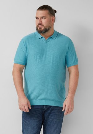 Homme avec barbe et cheveux attachés, portant un polo turquoise à manches courtes et un jean bleu, debout devant un fond gris uni.