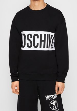 Persona che indossa una felpa nera con una larga striscia bianca e una grande scritta "MOSCHINO" sul petto, abbinata a pantaloni neri con il logo Moschino.