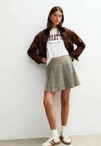 Chaqueta marrón de cuello sobre una camiseta gráfica blanca, combinada con una falda de cuadros gris con pliegues y zapatillas blancas con detalles marrones.