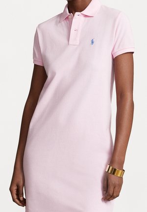 Sukienka polo w jasnoróżowym bawełnianym materiale, z kołnierzykiem, krótkimi rękawami i haftowanym niebieskim logo po lewej stronie klatki piersiowej.
