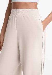 Helle beigefarbene Hose mit elastischem Bund, seitlichen Taschen und schwarzen Knopfakzenten entlang der Außennaht. Glattes, strukturiertes Material.