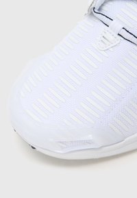 Biały but sportowy z teksturowaną górą z pionowymi wycięciami, gładką gumową podeszwą i cienkimi czarnymi akcentami.