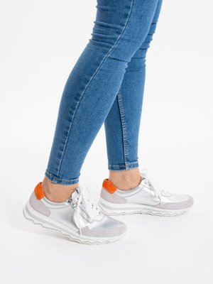 Person wearing blue skinny jeans and silver sneakers with white soles and orange heel accents, standing against a white background.