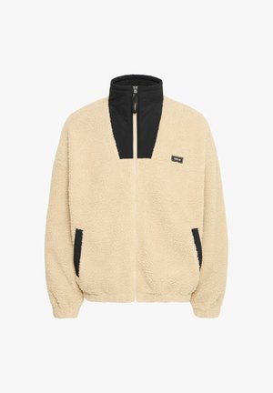 Beige fleece jacket with black nylon accents, high collar, full zip, and side pockets; texture appears soft and fluffy.