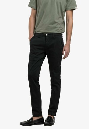 Hombre con pantalones negros ajustados, mocasines negros y una camiseta de manga corta verde oliva, de pie con las manos relajadas a los lados.