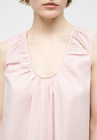 Γυναίκα που φοράει αμάνικη μπλούζα ανοιχτού ροζ χρώματος με σούρες στο ντεκολτέ και χρυσούς κρίκους στα αυτιά, εμφανιζόμενη από το κολάρο μέχρι τα χείλη.