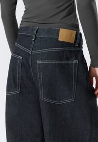 Mørk denimjeans med høyt liv, hvit søm, to baklommer og en brun skinnetikett i livet.