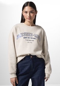 Felpa beige chiara con testo stampato blu, con scollo rotondo e vestibilità ampia. Abbinato a jeans a vita alta di colore blu scuro.