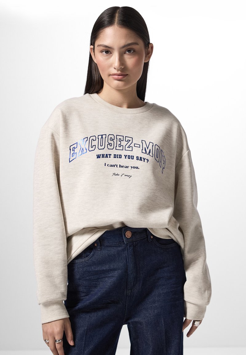 Felpa beige chiara con testo stampato blu, con scollo rotondo e vestibilità ampia. Abbinato a jeans a vita alta di colore blu scuro.