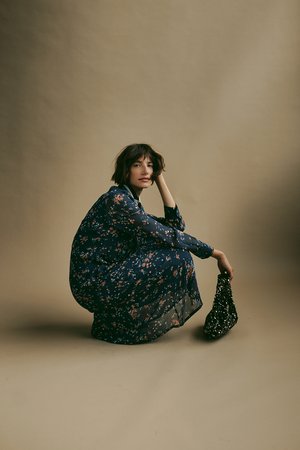 Navy bloemenjurk met lange mouwen en een lichte, doorschijnende textuur. Model houdt een sprankelende zwarte handtas vast terwijl ze gehurkt tegen een neutrale achtergrond staat.