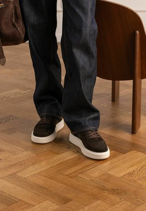 Persoon die donkere spijkerbroek en bruine sneakers met witte zolen draagt, staat op een houten parketvloer naast een houten stoel.