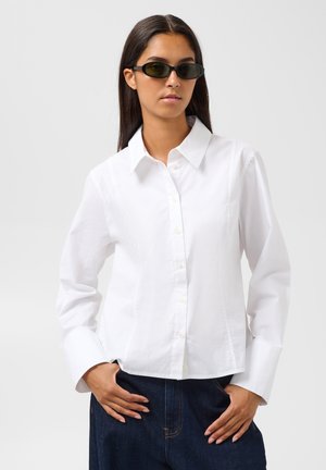 Femme aux longs cheveux foncés portant des lunettes de soleil noires, une chemise blanche boutonnée et un jean bleu foncé, posant avec les mains près des poches.