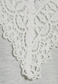 Dettagli in pizzo bianco su un tessuto grigio chiaro, con motivi floreali intricati e una finitura testurizzata, che arricchiscono il design del capo.