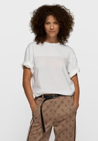 Camiseta blanca de gran tamaño con mangas enrolladas, combinada con pantalones marrones con estampado, cinturón negro y franjas laterales.