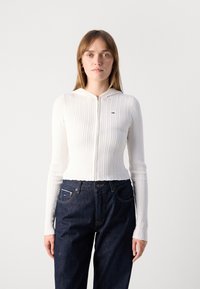 Maglione bianco a coste con un design aderente e chiusura con zip, dotato di cappuccio. Abbinato a jeans di denim scuri. Branding minimale sul petto.