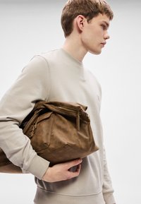 Καφέ δερμάτινη τσάντα με υφή, κλείσιμο με φερμουάρ και πλαϊνή τσέπη· φορεμένη στον ώμο από άτομο που φοράει ανοιχτό γκρι φούτερ.