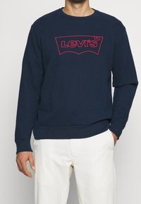 Man i marinblå Levi's sweatshirt med röd logotyp och vita byxor, stående med ena handen i fickan mot en enkel bakgrund.