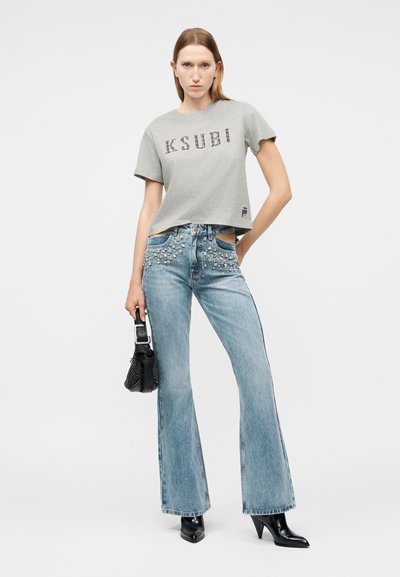 Γυναίκα που φοράει γκρι t-shirt Ksubi, τζιν με ανοίγματα σε ανοιχτό μπλε χρώμα με τρουκς, μαύρες μυτερές μπότες, κρατώντας μια μαύρη τσάντα με ασημένιες τρούκς.