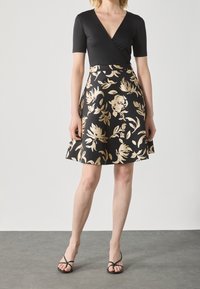 Svart wrap-top med korta ärmar, tillsammans med en flippskjol med ett guldblommigt mönster mot en svart bakgrund. Svarta sandaler med fyrkantig tå.