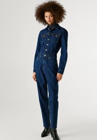 Džínový overál v tmavo-modrej farbe. Má predné gombíkové zapínanie, vrecká na hrudi a dlhé rukávy. Priliehavý strih s rovnými nohavicami. Hladká textúra.