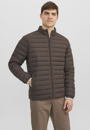 Giubbotto imbottito marrone con design trapuntato verticale, colletto alto e zipper frontale. Caratterizzato da polsini elasticizzati e tasche laterali, indossato sopra un tessuto beige.