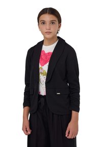 Blazer corto nero con maniche lunghe e colletto sciallato, dotato di una tasca frontale e tessuto a trama, indossato sopra una maglietta bianca con grafica.