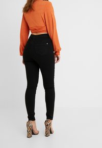 Jeans skinny negros combinados con una blusa de manga larga acanalada naranja. Unos zapatos de tacón con estampado de serpiente completan el look. Se muestra la vista desde atrás.