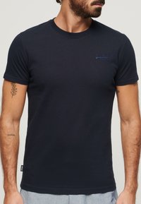Mand iført en ensfarvet mørk marineblå T-shirt med korte ærmer og et lille broderet logo på brystet, samtidig med at hans tatoverede arme er synlige.