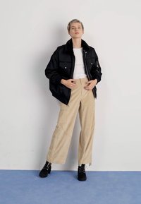 Sort bomberjakke med kunstpelskrave, hvid t-shirt, beige vidde ben bukser med lommer, sorte snørebåndssko, der står op ad en neutral baggrund.