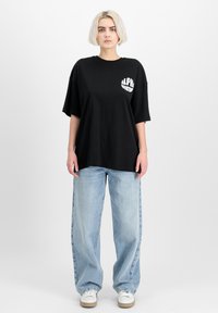 Černé oversized tričko s bílým logem "ALPHA INDUSTRY" na levé hrudi, spárované s světle modrými širokými džíny a bílými teniskami.
