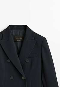 Blazer croisé bleu marine avec revers à encoche, boutons sur le devant et poche poitrine, présenté sur fond blanc.