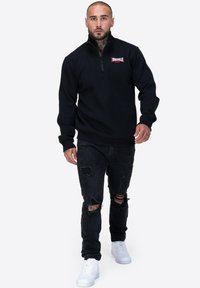 Pullover nero con zip a un quarto e logo rosso, abbinato a jeans neri strappati e sneakers bianche. Caratterizzato da maniche lunghe e polsini a coste.