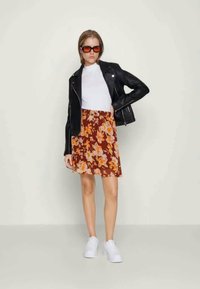 Casaco de motociclista em couro preto, t-shirt branca, saia floral castanha com detalhes laranja e amarelo, ténis brancos e óculos de sol.