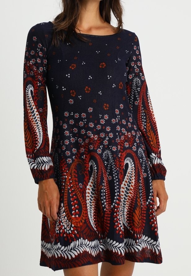 Femme portant une robe à manches longues bleu marine avec des motifs cachemire et floraux rouges, blancs et marron, debout les bras détendus.