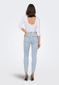 Γυναίκα που στέκεται με το χέρι στην πίσω τσέπη, φορώντας λευκή μπλούζα με ανοιχτή πλάτη, ανοιχτόχρωμα στενά τζιν και μπεζ ψηλοτάκουνα, σε απλό φόντο.