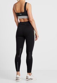 Leggings de sport noirs avec des panneaux en mesh, associés à un soutien-gorge de sport noir et gris. Les chaussures sont blanches avec des accents gris. Design minimaliste.