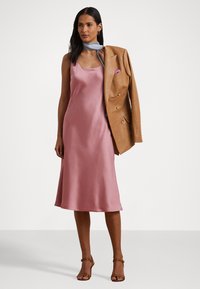 Vestido de cetim rosa, com corpete justo e saia ampla, complementado com um blaser de couro bege e um cachecol listrado azul ao pescoço.