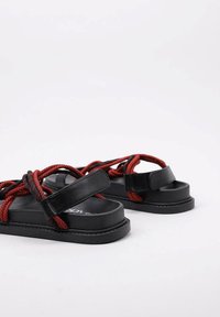 Sandalias negras con suela plana, con correas tejidas rojas. Las correas están entrelazadas con una textura similar al ante.