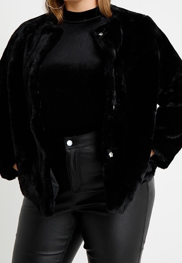 Frau, die eine schwarze Kunstpelzjacke über einem schwarzen Rollkragenpullover und einer hochtaillierten schwarzen Lederhose trägt, steht mit den Händen in den Taschen.