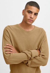 Un pull beige texturé avec un col rond et des poignets côtelés. Le mannequin a les bras croisés, mettant en valeur le tissage et l'ajustement du tissu.