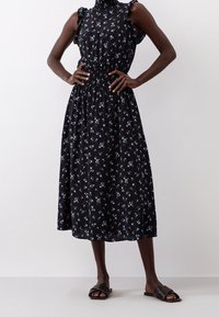 Robe midi noire à motifs floraux bleus, avec des détails froncés aux épaules et une taille élastiquée. Portée avec des sandales plates noires.