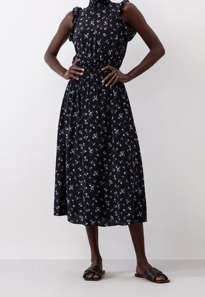 Robe midi noire à motifs floraux bleus, avec des détails froncés aux épaules et une taille élastiquée. Portée avec des sandales plates noires.