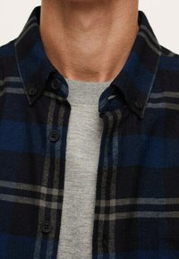 Camicia di flanella blu scuro con motivo a quadri neri e grigi, dotata di colletto button-down e bottoni coordinati, indossata sopra una maglietta grigia.