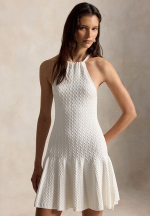 Γυναίκα με μακριά σκούρα μαλλιά που φοράει λευκό μίνι φόρεμα αμάνικο με πλεχτή λεπτομέρεια, λαιμόκοψη halter και φουσκωτή φούστα, σε απλό φόντο.