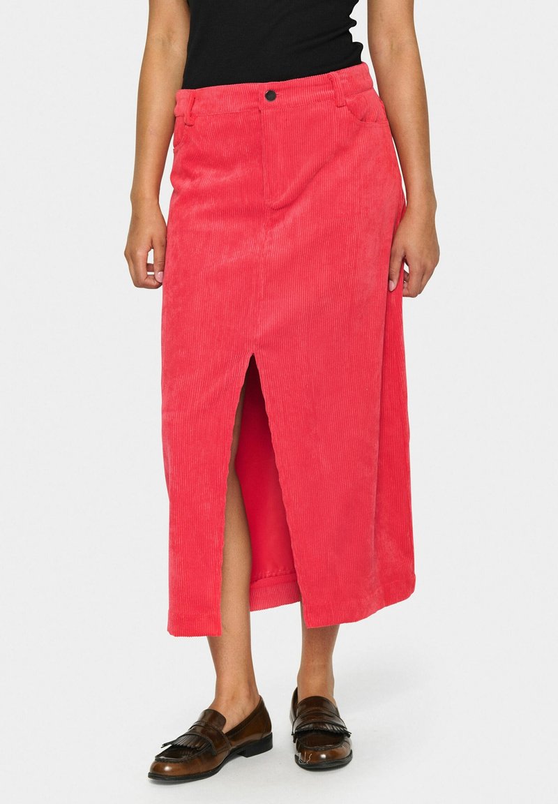 Femme portant une jupe midi en velours côtelé rouge avec une fente à l’avant, un haut noir et des mocassins en cuir marron, debout devant un fond uni.