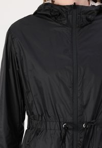 Casaco leve preto com fecho de correr à frente, cordão na cintura e capuz. Apresenta um tecido liso e brilhante e punhos elásticos.